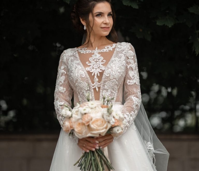 Bride in a lace wedding gown with a sheer neckline, holding a blush peach and ivory bouquet outdoors among dark green foliage.