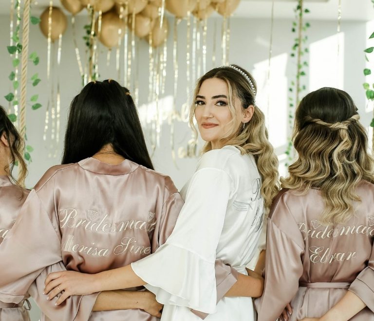 Bride in a white robe smiles as her bridesmaids in blush satin robes hug her, backs showing 'Bridesmaid' text, with gold balloons overhead.